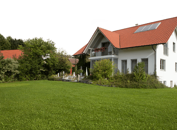 Ein weißes Einfamilienhaus mit rotem Dach und Solarpanel steht in einem großen Garten mit Rasen und Büschen.